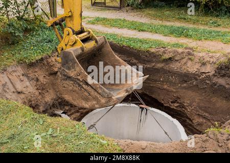 Pelle hydraulique avec un godet, s'abaissant dans la fosse sur des câbles en acier anneau d'égout en béton. Construction ou réparation d'un égout. Banque D'Images