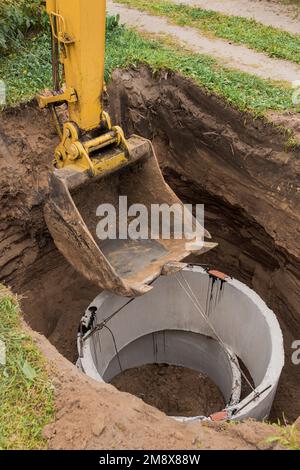 Pelle hydraulique avec un godet, s'abaissant dans la fosse sur des câbles en acier anneau d'égout en béton. Construction ou réparation d'un égout. Banque D'Images