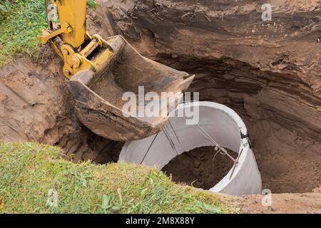 Pelle hydraulique avec un godet, s'abaissant dans la fosse sur des câbles en acier anneau d'égout en béton. Construction ou réparation d'un égout. Banque D'Images