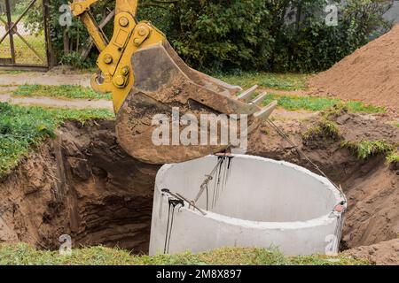 Pelle hydraulique avec un godet, s'abaissant dans la fosse sur des câbles en acier anneau d'égout en béton. Construction ou réparation d'un égout. Banque D'Images