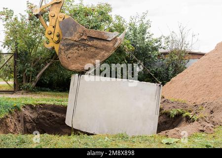 Pelle hydraulique avec un godet, s'abaissant dans la fosse sur des câbles en acier anneau d'égout en béton. Construction ou réparation d'un égout. Banque D'Images