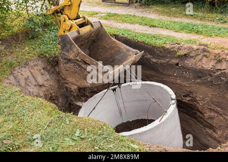 Pelle hydraulique avec un godet, s'abaissant dans la fosse sur des câbles en acier anneau d'égout en béton. Construction ou réparation d'un égout. Banque D'Images