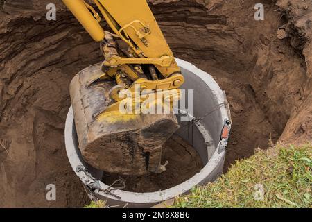 Pelle hydraulique avec un godet, s'abaissant dans la fosse sur des câbles en acier anneau d'égout en béton. Construction ou réparation d'un égout. Banque D'Images