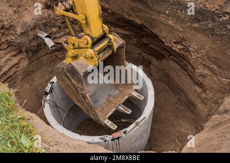 Pelle hydraulique avec un godet, s'abaissant dans la fosse sur des câbles en acier anneau d'égout en béton. Construction ou réparation d'un égout. Banque D'Images