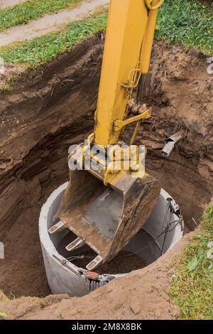 Pelle hydraulique avec un godet, s'abaissant dans la fosse sur des câbles en acier anneau d'égout en béton. Construction ou réparation d'un égout. Banque D'Images