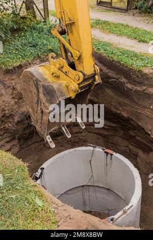 Pelle hydraulique avec un godet, s'abaissant dans la fosse sur des câbles en acier anneau d'égout en béton. Construction ou réparation d'un égout. Banque D'Images