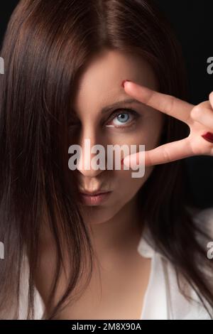 Portrait d'une femme adulte sérieuse tenant deux doigts près de l'œil, montrant un geste de victoire ou de paix et regardant l'appareil photo. Un beau brunette Banque D'Images