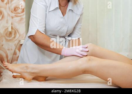 une esthéticienne conduit une procédure d'épilation du sucre avec du miel sur les jambes d'une fille couchée dans un salon professionnel de la santé et des soins de la peau. Mod Banque D'Images