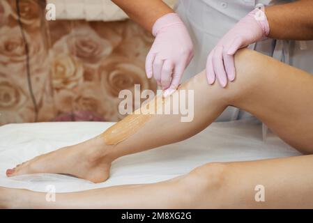 une esthéticienne conduit une procédure d'épilation du sucre avec du miel sur les jambes d'une fille couchée dans un salon professionnel de la santé et des soins de la peau. Mod Banque D'Images