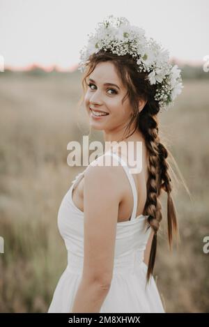 portrait d'une fille souriante heureuse avec des tresses et une couronne fleurie en robe blanche de style boho en été à l'extérieur. Ajout d'une simulation de petit grain du film p Banque D'Images