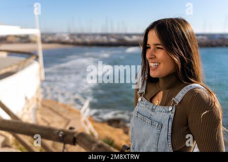 Vue latérale d'une jeune femme gaie en col roulé brun et en denim dans l'ensemble souriant avec joie en se tenant debout sur un remblai contre la mer pendant les vacances Banque D'Images