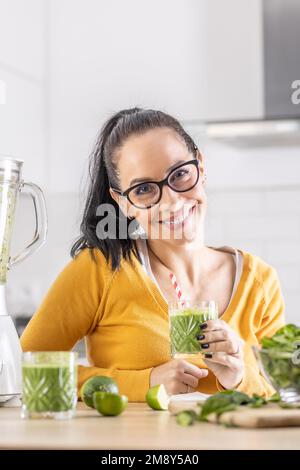 Bonne femme avec un smoothie aux épinards ou un shake végétarien assis dans sa cuisine. Banque D'Images