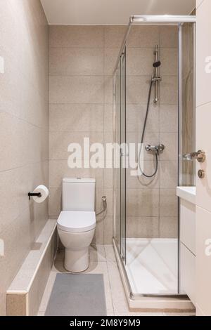 Entrée à une petite salle de bains avec carrelage en marbre beige sur les murs et le sol, toilettes blanches propres, lavabo, douche à parois de verre. Salle de bains est Banque D'Images