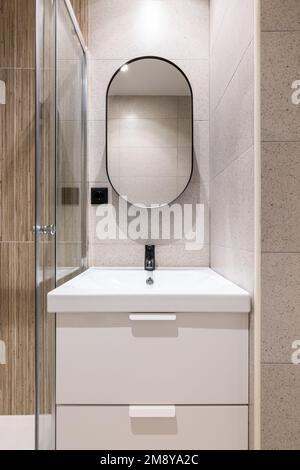 Vue rapprochée d'une alcôve de salle de bains avec éclairage artificiel lumineux, avec un lavabo blanc en céramique solide sur un meuble-lavabo à deux tiroirs. Grand miroir ovale moderne sur le Banque D'Images