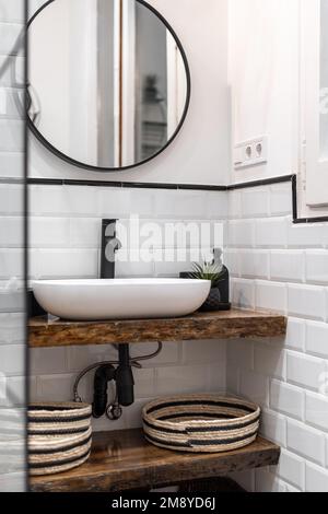Salle de bains avec murs en briques blanches, lavabo ovale sur un dessus en marbre de style bois, miroir rond sur un mur à cadres sombres qui reflète une porte. Céramique noire Banque D'Images