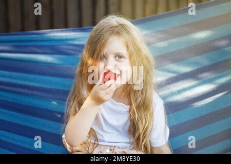 une petite fille blonde aux cheveux longs mange une fraise dans le jardin assis dans un hamac. Banque D'Images