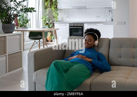 Bonne femme afro-américaine décontractée dans un casque qui ferme les yeux en écoutant de la musique assis sur un canapé Banque D'Images