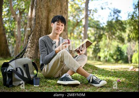 Beau et heureux jeune asiatique étudiant d'université de sexe masculin se trouve sous l'arbre dans le parc, manger de la pomme tout en lisant un livre. concept loisir et passe-temps Banque D'Images