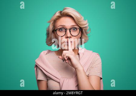 La jeune femme appuie l'index sur les lèvres semble mystérieusement sur l'appareil photo faisant le signe shh geste isolé sur le mur vert de studio. Banque D'Images