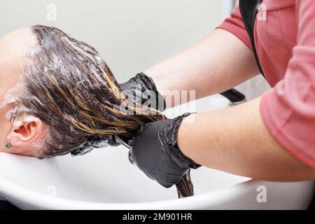 Le coiffeur en gants noirs lavant les cheveux de la femme brunette dans le salon de beauté Banque D'Images