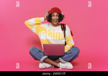 Jeune homme des caraïbes en casque riant et en utilisant un ordinateur portable isolé sur fond rose Banque D'Images