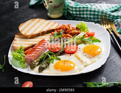 Petit déjeuner Keto. Petit déjeuner. Œufs frits, pain grillé, asperges vertes, haricots et saucisses sur une assiette blanche. Banque D'Images