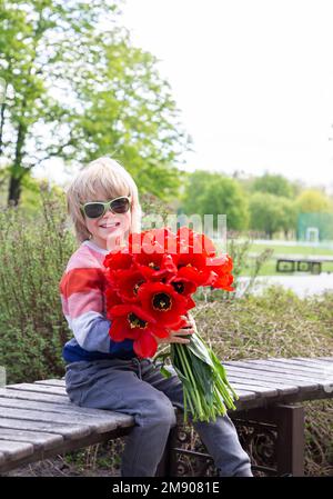 élégant garçon de 6 ans tient un grand bouquet de tulipes rouges dans ses mains. Joyeux enfant souriant avec des fleurs. Fête des mères, Saint-Valentin, Banque D'Images