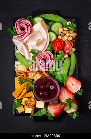 Assiette d'antipasti : jambon en tranches, salami, crackers, fraises, légumes et plateau de fromages à bord sur fond sombre. Table de hors-d'œuvre avec Banque D'Images