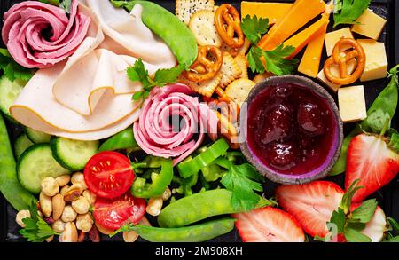 Assiette d'antipasti : jambon en tranches, salami, crackers, fraises, légumes et plateau de fromages à bord sur fond sombre. Table de hors-d'œuvre avec Banque D'Images
