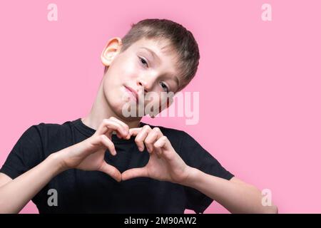 Un adolescent en T-shirt noir donne une forme de coeur avec ses mains et sourit sur fond rose. Amour, relations. saint valentin. Banque D'Images