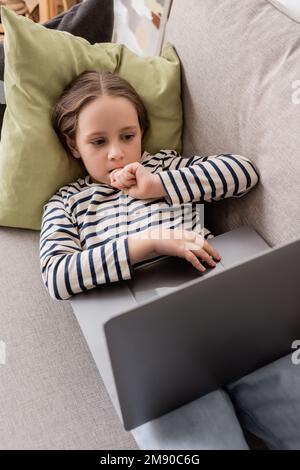 vue à grand angle de la préadolescence ciblée fille dans des vêtements décontractés en utilisant un ordinateur portable tout en étant allongé sur un canapé, image de stock Banque D'Images