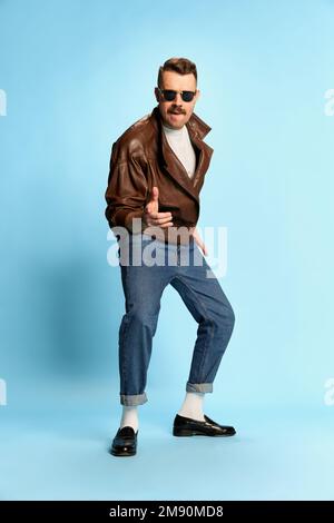 Portrait d'homme brutal et beau en Jean, veste en cuir et lunettes de soleil posant, dansant sur fond bleu studio. Confiance. Concept des émotions Banque D'Images