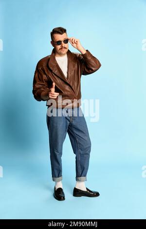 Charismatique. Portrait d'un homme brutal et beau en Jean, veste en cuir et lunettes de soleil posant sur fond bleu studio. Concept des émotions Banque D'Images