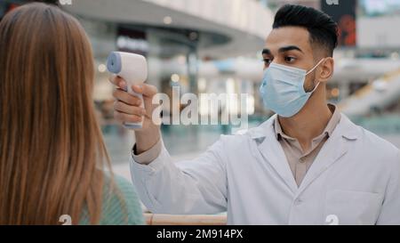 Médecin homme ethnique Indien travailleur médical visage dans le masque blouse de laboratoire uniforme arrêter méconnaissable femme mesure la température de dépistage fièvre se penchant à Banque D'Images