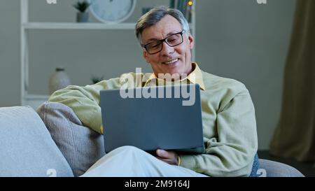 Grand-père heureux s'asseoir sur le canapé salon âge adulte homme caucasien regardant l'écran de travail d'ordinateur portable navigue à distance des nouvelles sur Internet faire en ligne Banque D'Images