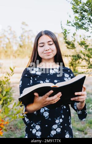 Jeune fille religieuse souriante lisant sa bible, à l'extérieur dans le champ au coucher du soleil. Renouveau spirituel. Banque D'Images