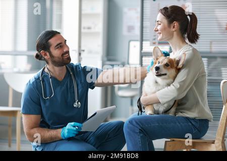 Joyeux jeune vétérinaire en gants et uniforme consultant femme propriétaire de chien corgi gallois de race pure au bureau médical Banque D'Images