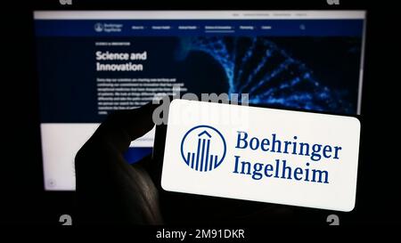 Personne tenant le téléphone portable avec le logo de la société pharmaceutique Boehringer Ingelheim sur l'écran en face de la page web d'affaires. Mise au point sur l'affichage du téléphone. Banque D'Images