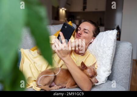 Jeune femme allongé sur un canapé avec son petit chien utilisant un téléphone mobile et un casque prenant selfie avoir du plaisir grimaquant au téléphone mobile. Banque D'Images
