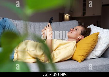 Femme pensive avec un casque utilisant son téléphone portable tout en étant allongé sur un canapé dans le salon à la maison. Banque D'Images