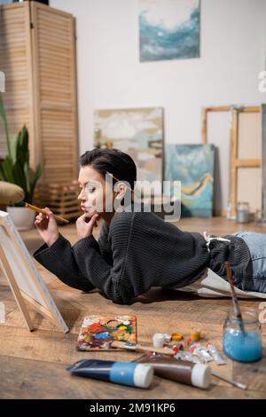 Un jeune artiste se panse sur toile tout en étant allongé sur le sol dans un atelier Banque D'Images