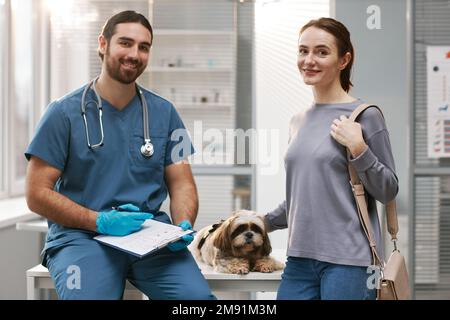 Jeune femme propriétaire du terrier du Yorkshire mignon, son animal de compagnie et clinicien vétérinaire en uniforme bleu regardant la caméra dans le bureau médical Banque D'Images