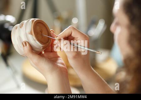 Jeune femme faisant de la poterie à l'intérieur dans un studio créatif artisan concept assis à table à l'aide de l'outil de modélisation créant des détails sur la tasse. Banque D'Images