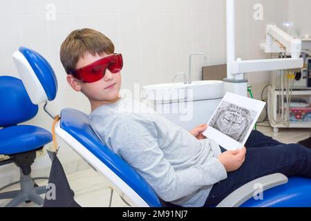 Un garçon heureux dans des lunettes de sécurité avec des rayons X dentaires (radiographies) dans ses mains assis dans un cabinet de dentiste Banque D'Images