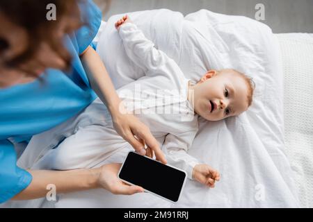 vue de dessus du petit enfant dans la barboteuse allongé sur le lit d'hôpital près de maman floue tenant un smartphone avec écran blanc, image de stock Banque D'Images