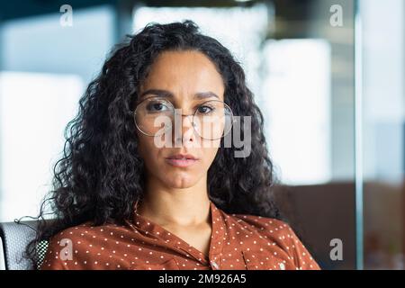 Portrait en gros plan d'une femme d'affaires confiante et sérieuse, une femme hispanique portant des lunettes à l'intérieur du bureau regardant l'appareil photo avec des cheveux bouclés. Banque D'Images