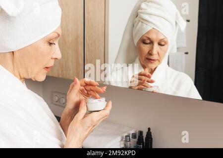 Femme âgée avec une serviette enveloppée sur une tête tenant une crème. Femme âgée se préparant à appliquer un hydratant dans la salle de bain. Banque D'Images