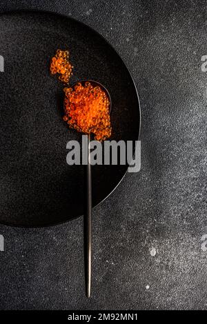 Cuillère noire pleine de caviar rouge sur plaque noire et fond de table en béton de pierre de même couleur Banque D'Images
