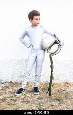 Corps complet de garçon en costume d'espace argenté debout sur un sol jaune herbacé avec un casque à la main et regardant loin Banque D'Images