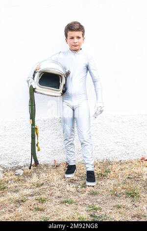 Sur toute la longueur d'un garçon de la préadolescence enfané dans un costume d'espace debout sur une herbe garée près d'un mur de plâtre blanc et tenant un casque Banque D'Images